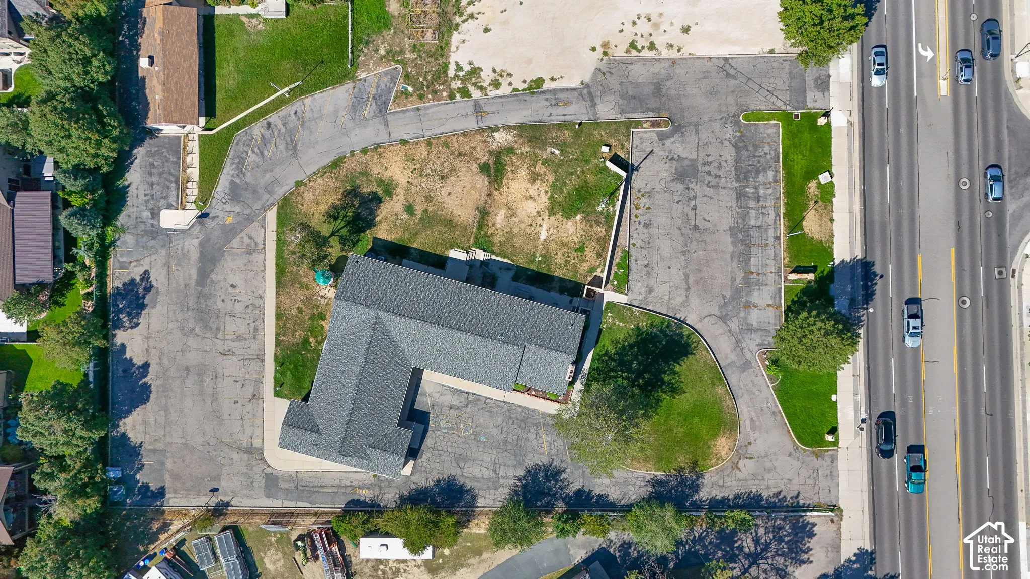 Asphalt parking areas surrounding the building, L-shaped building with a dark gray shingle roof, adjacent to a multi-lane roadway, areas of established lawn and mature trees