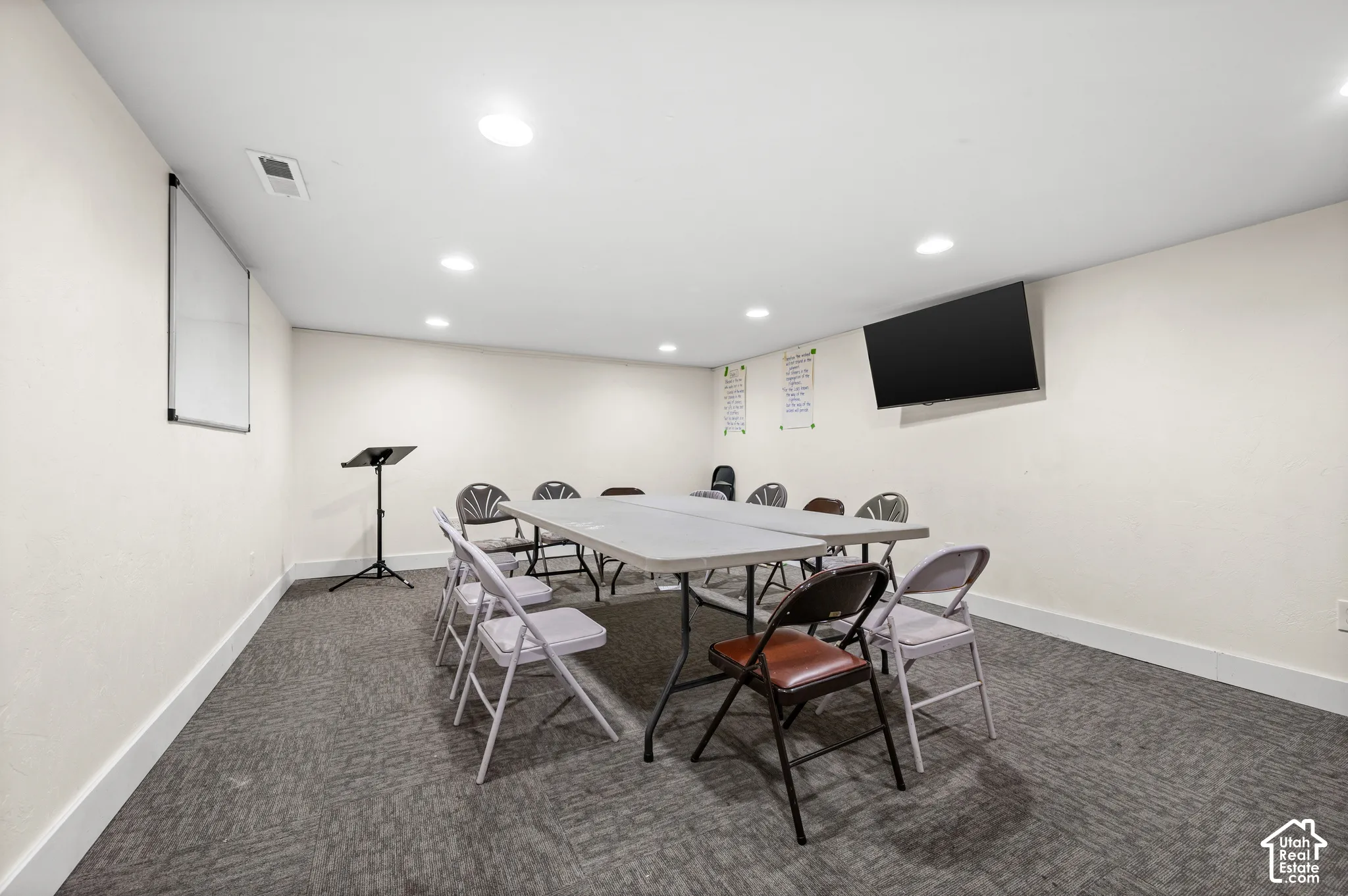Recessed lighting illuminates a spacious room featuring a mounted display, whiteboard, and grey patterned carpeting