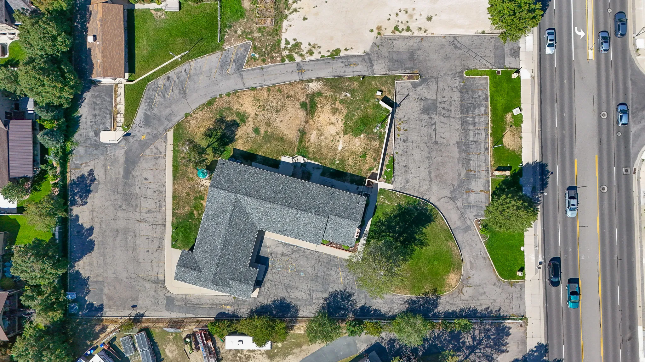 Asphalt parking areas surrounding the building, L-shaped building with a dark gray shingle roof, adjacent to a multi-lane roadway, areas of established lawn and mature trees