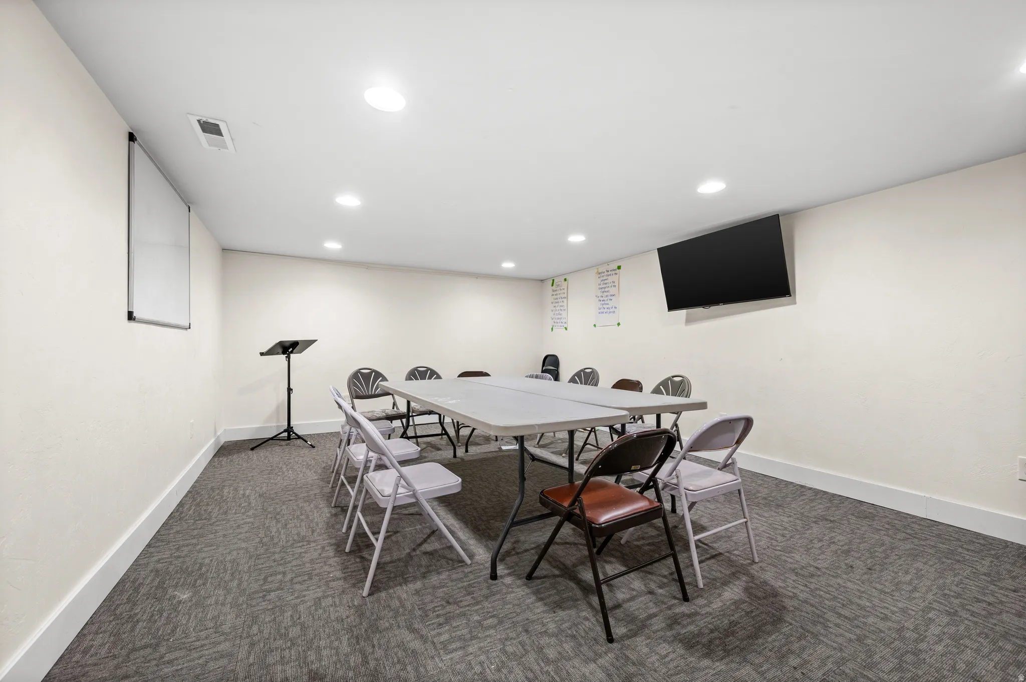 Recessed lighting illuminates a spacious room featuring a mounted display, whiteboard, and grey patterned carpeting