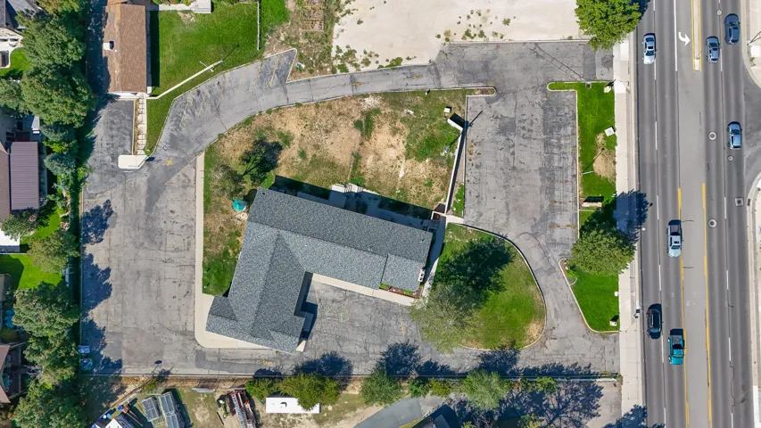 Asphalt parking areas surrounding the building, L-shaped building with a dark gray shingle roof, adjacent to a multi-lane roadway, areas of established lawn and mature trees