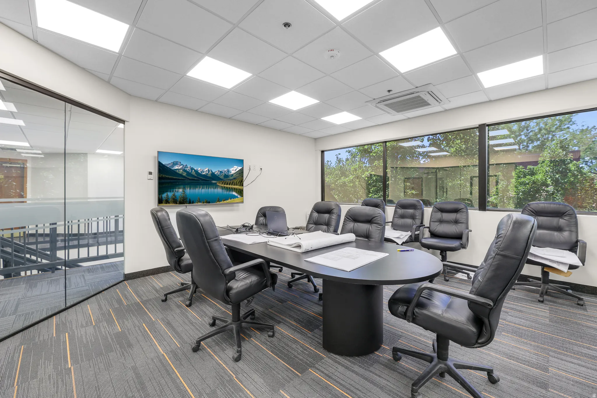Conference room with a large wall-mounted display, full-height exterior windows, suspended ceiling with recessed lighting, and patterned carpet flooring