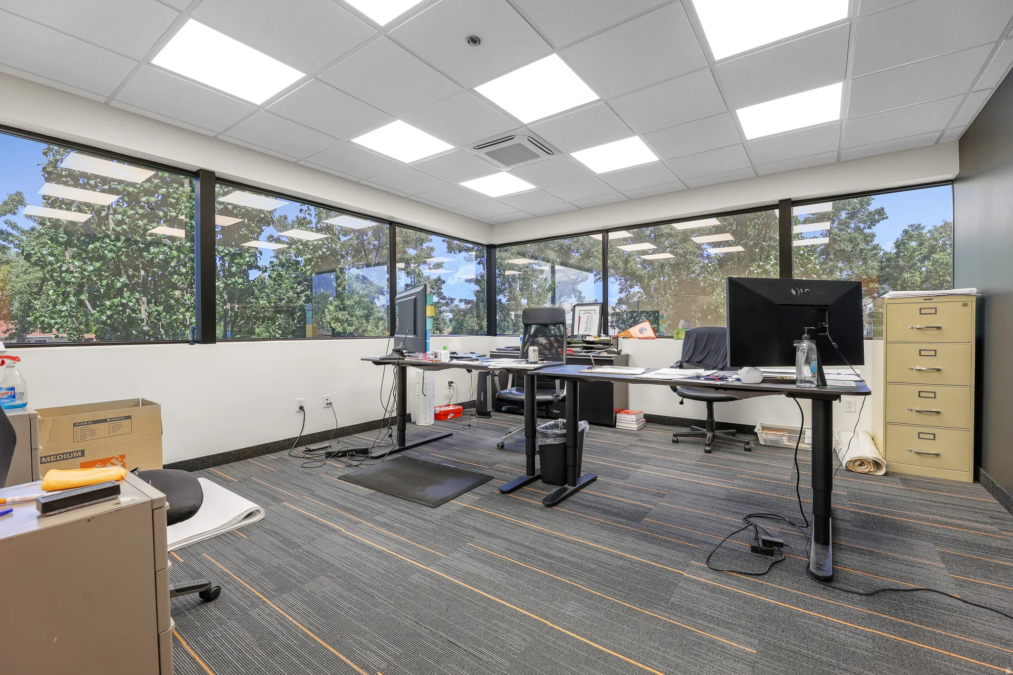 Expansive window-lined walls, commercial-grade ceiling tiles with integrated lighting, a ceiling-mounted HVAC register, and patterned carpet flooring