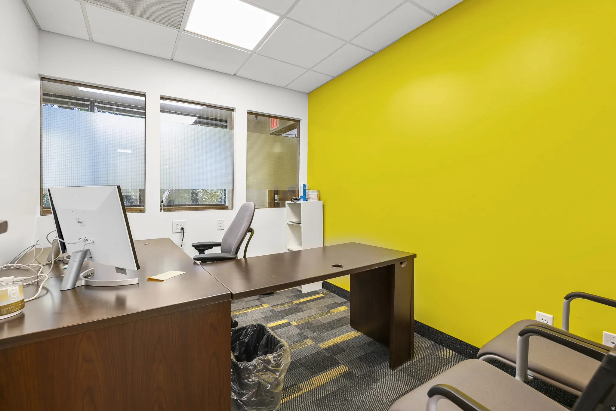 Frosted window panels, acoustical tile ceiling, and a vibrant yellow accent wall complement the modular wood-finish desk