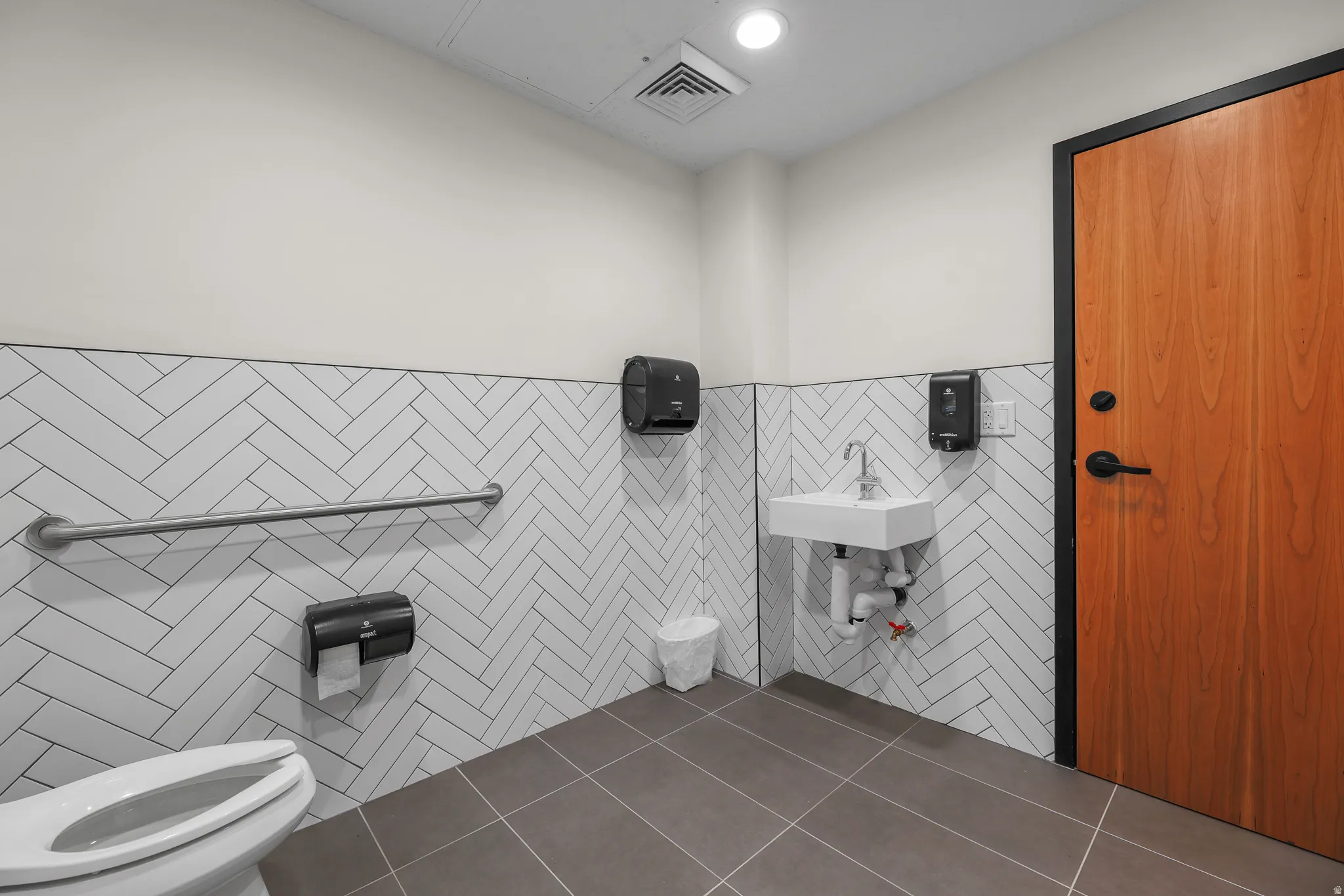 White subway tile with herringbone pattern, dark grey floor tiles, wood-finish door with black trim and hardware, wall-mounted sink, and a grab bar