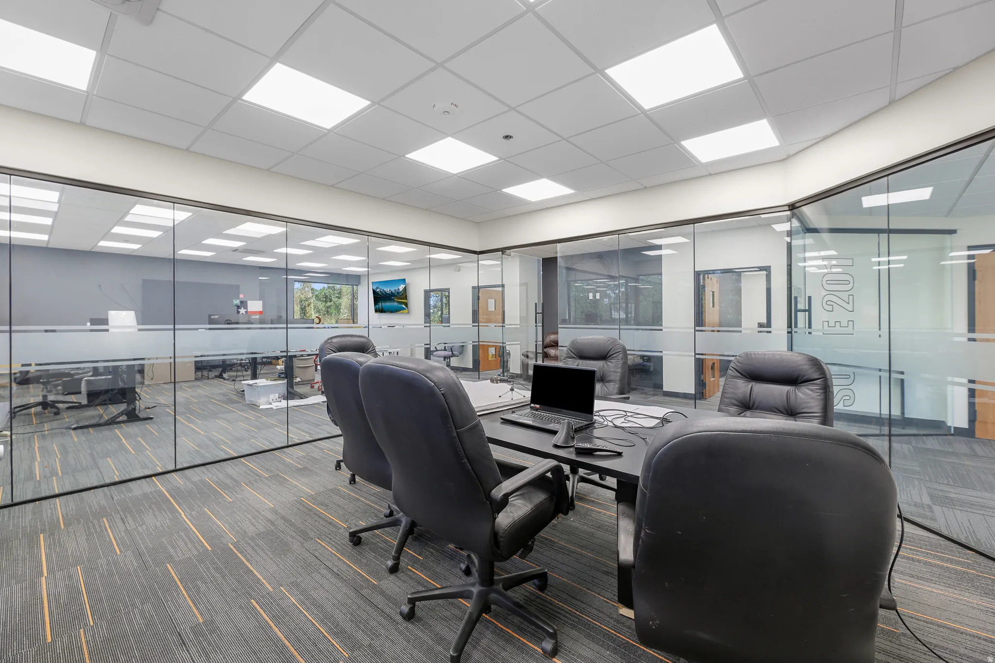 Glass-enclosed office space with carpet flooring, suspended ceiling with recessed lighting, and a wall-mounted display