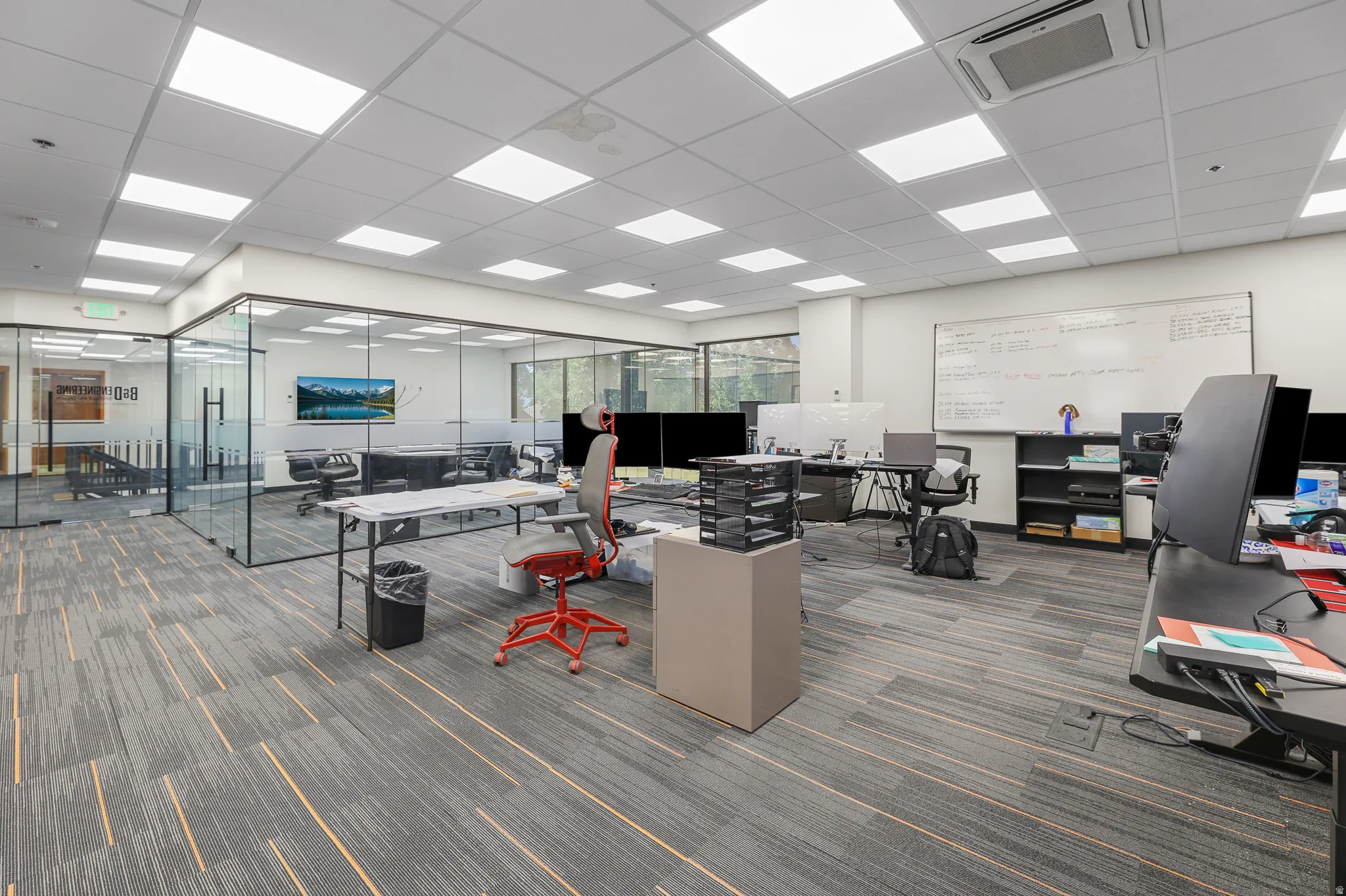Open-concept office space featuring modular carpet flooring, extensive recessed lighting, a glass-enclosed conference room, and a built-in whiteboard