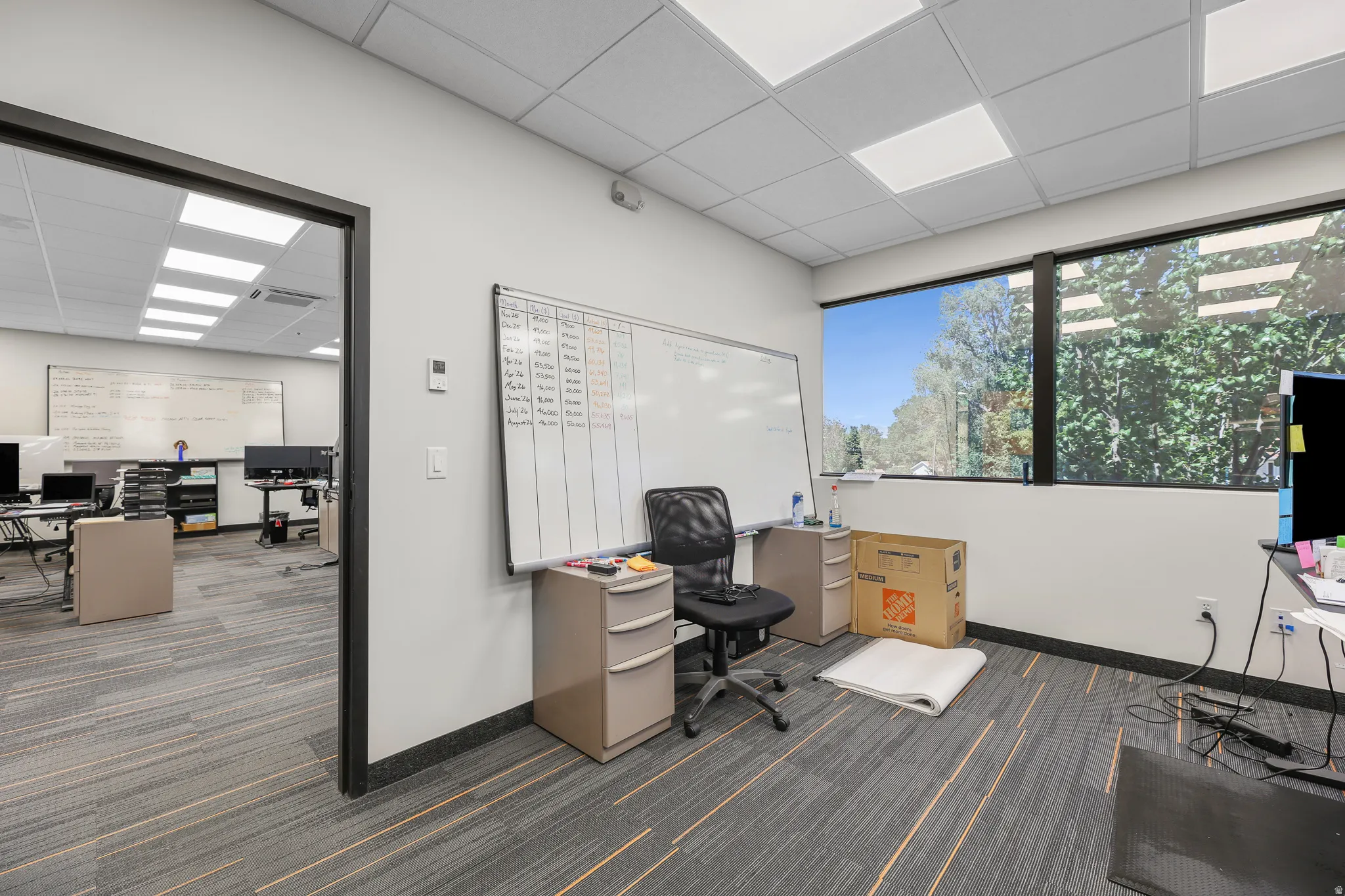 Expansive windows, drop ceiling with recessed lighting, wall-mounted whiteboard, and carpet tile flooring