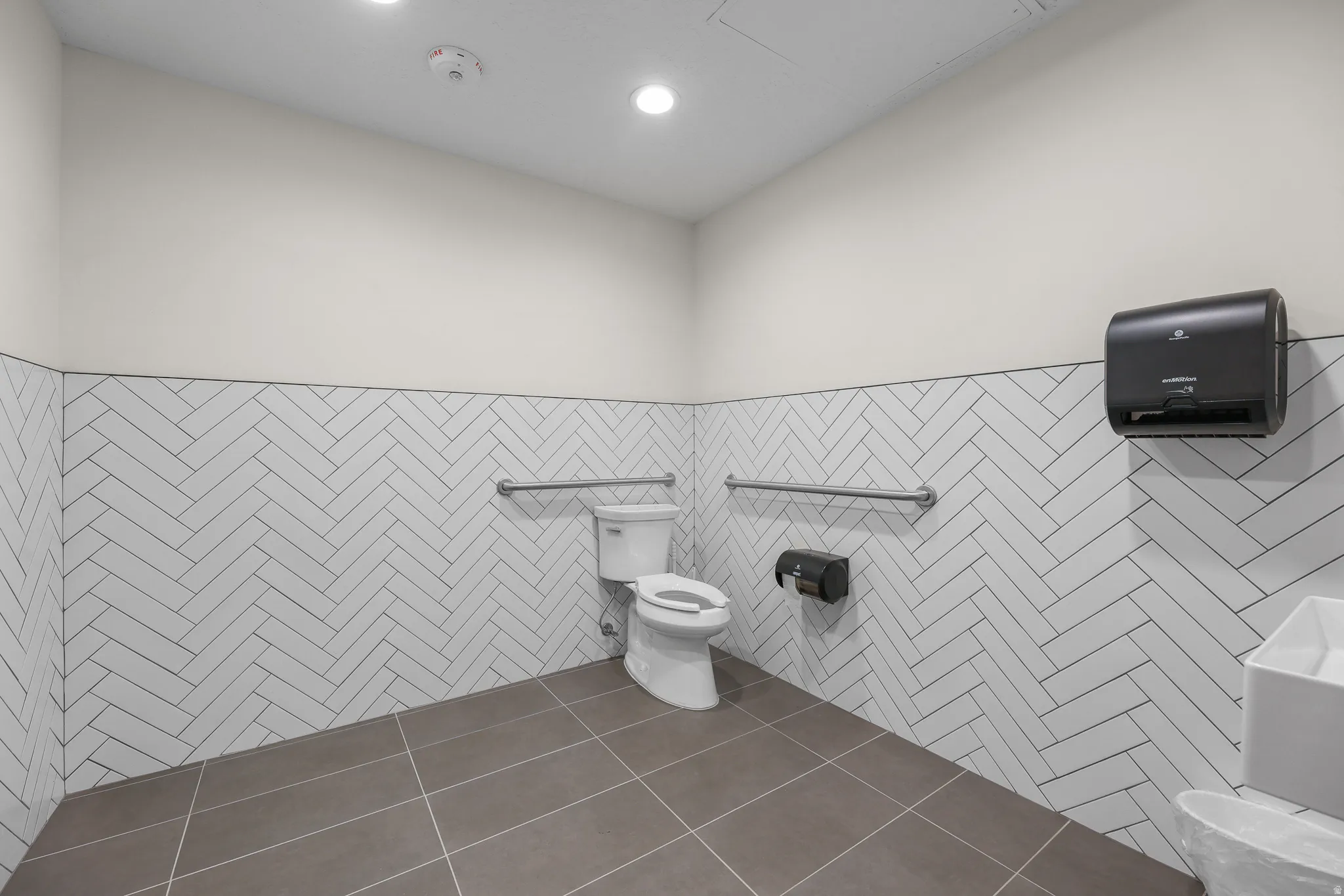 Herringbone tile wainscoting, recessed lighting, wall-mounted paper towel dispenser, stainless steel grab bars, and large format floor tiling