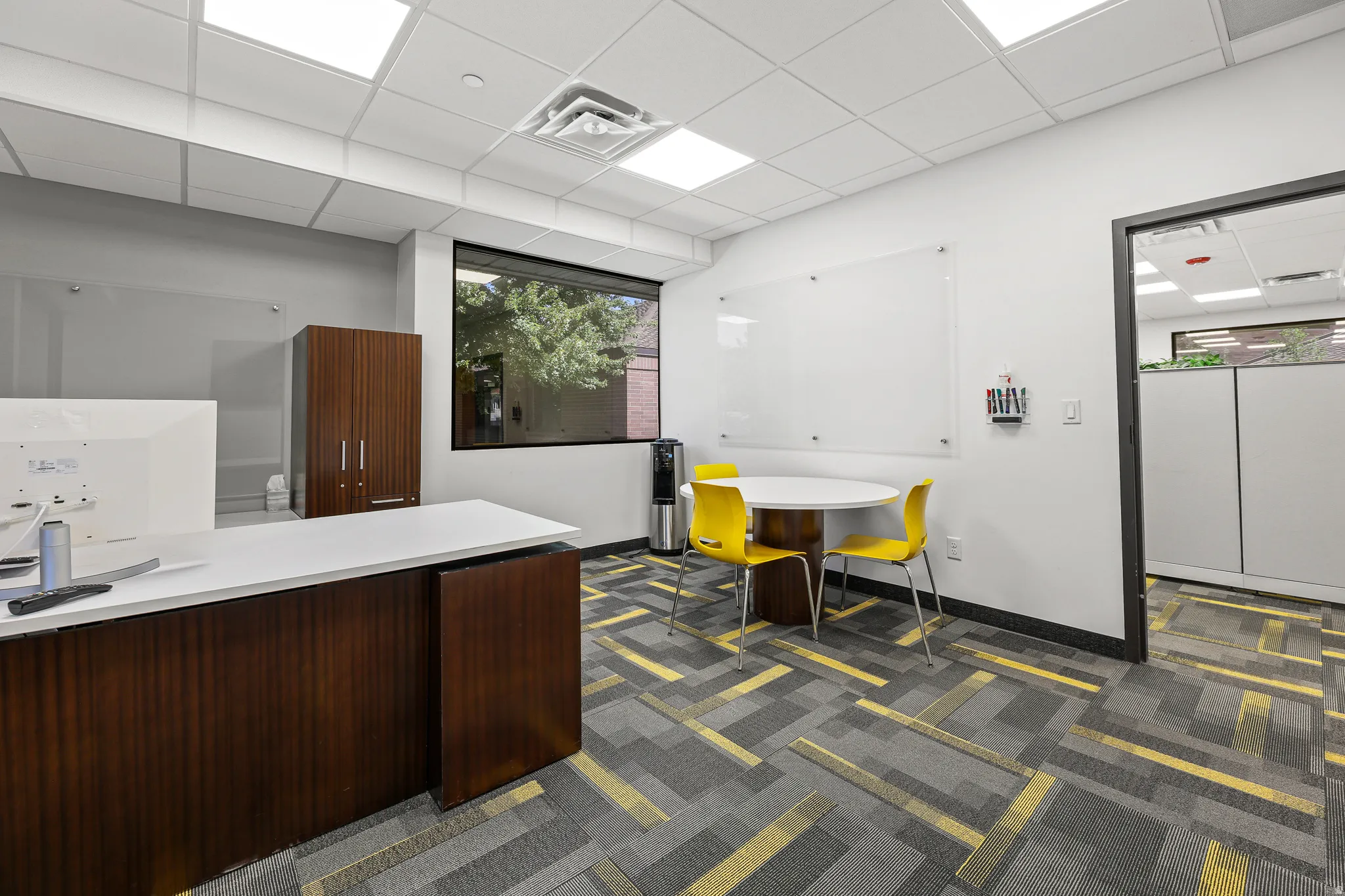 Custom-built desk with a white countertop and wood-finish base, ample natural light from a large window, patterned carpeting, and a drop ceiling with recessed lighting