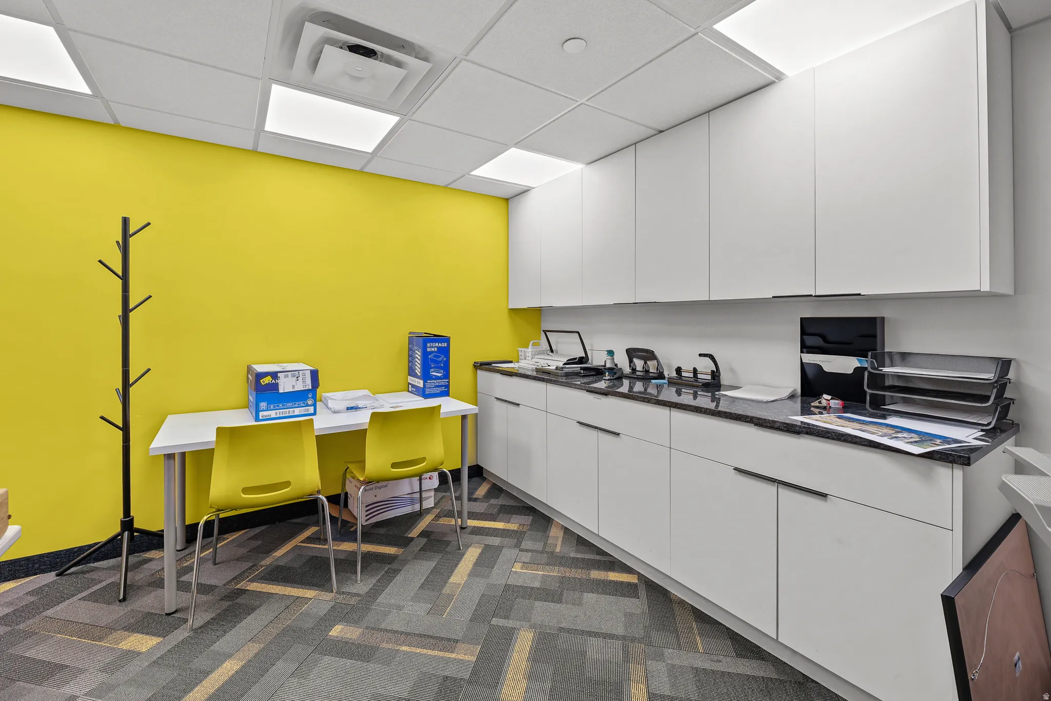 Contemporary office space featuring modular ceiling tiles with integrated lighting, a bright accent wall, extensive built-in white cabinetry, and patterned carpet flooring