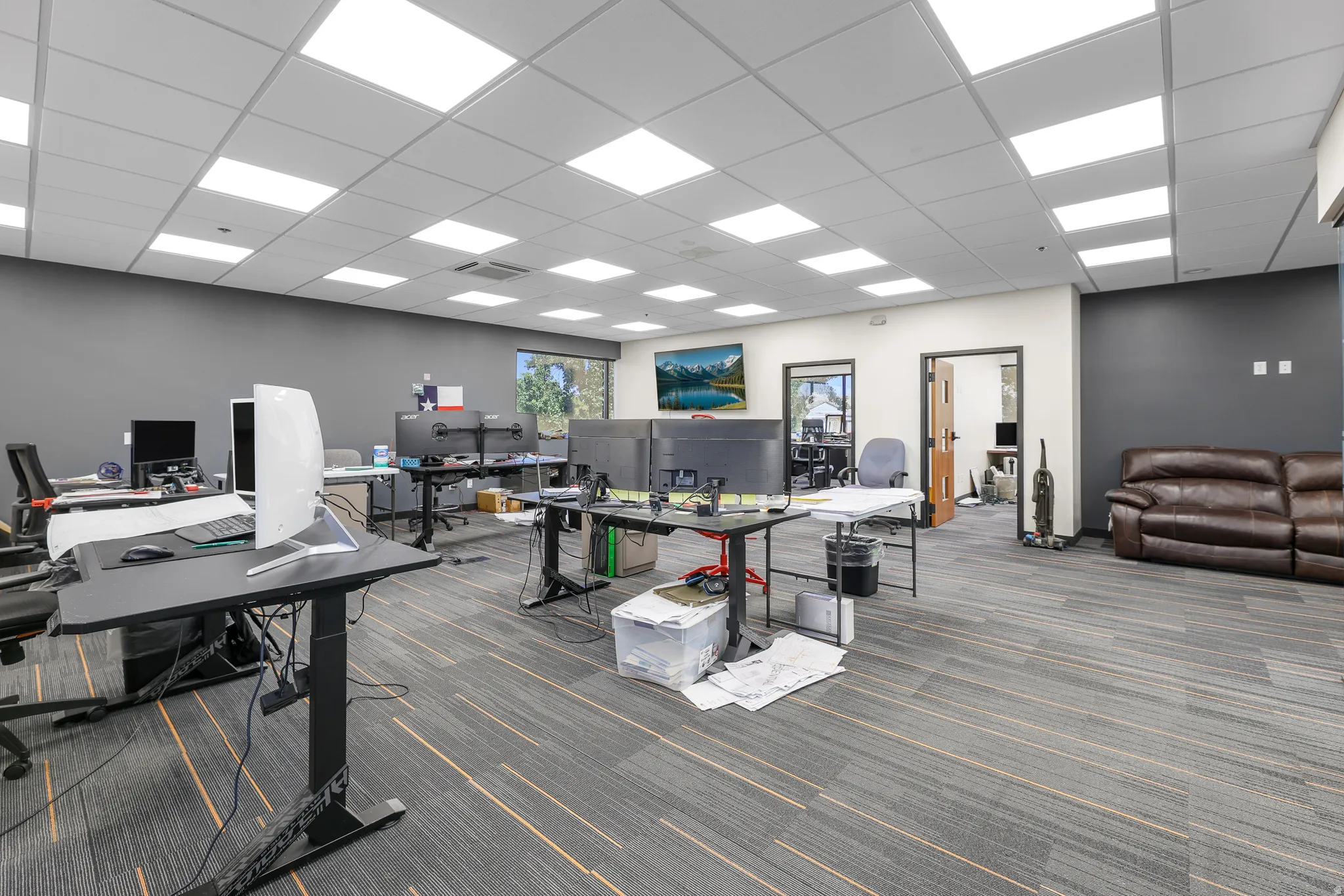 Open-plan office space featuring carpeted flooring with linear accents, suspended acoustical ceiling, recessed lighting, and a large window