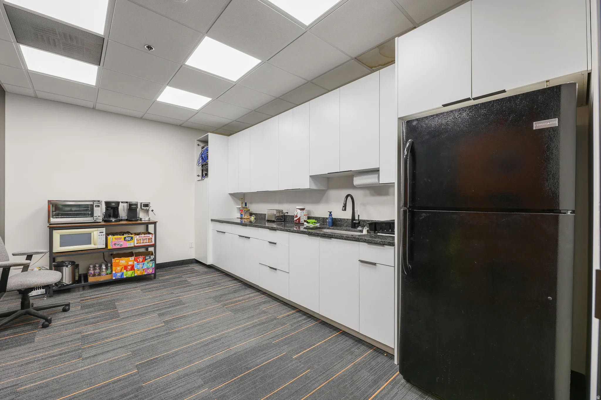 Kitchenette with white flat-panel cabinetry, dark speckled countertops, and a black refrigerator