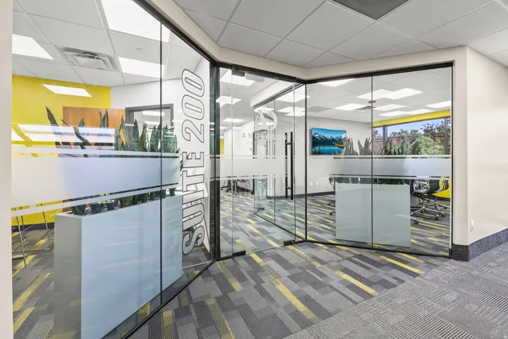 Glass-enclosed office space featuring a frosted entry door with suite number, acoustic ceiling tiles with recessed lighting, patterned carpet flooring, and a window with exterior views