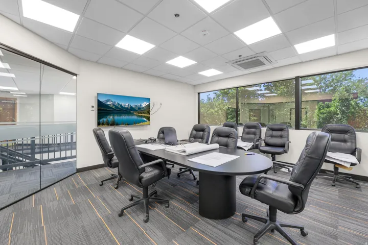 Conference room with a large wall-mounted display, full-height exterior windows, suspended ceiling with recessed lighting, and patterned carpet flooring
