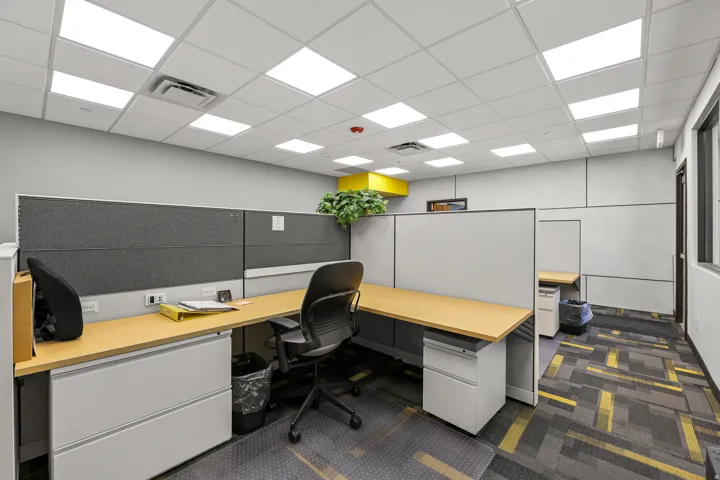 Modern office workspace featuring dual workstations with yellow wood-finish desktops and gray fabric privacy panels, ergonomic black task seating, white storage cabinetry with integrated shelving, geometric patterned carpet flooring, and bright LED recess