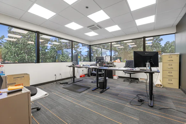Expansive window-lined walls, commercial-grade ceiling tiles with integrated lighting, a ceiling-mounted HVAC register, and patterned carpet flooring