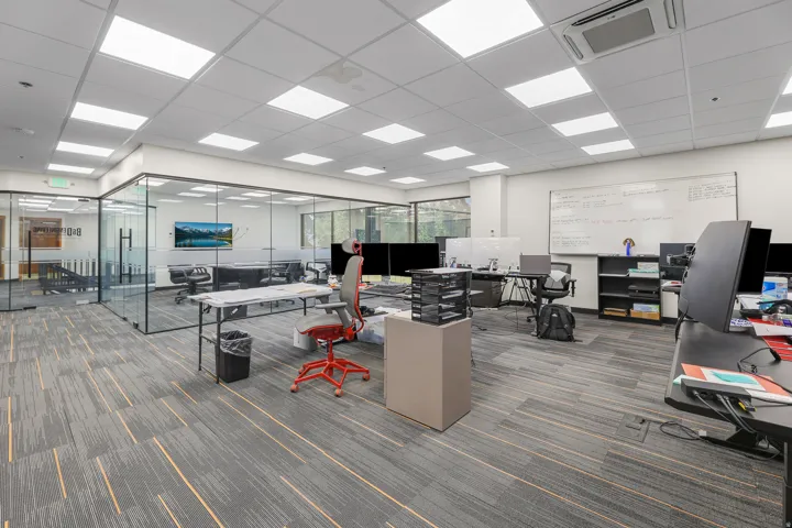 Open-concept office space featuring modular carpet flooring, extensive recessed lighting, a glass-enclosed conference room, and a built-in whiteboard