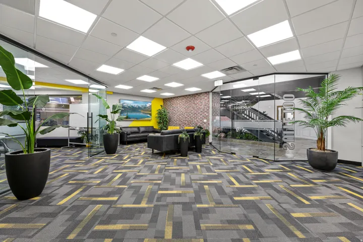 Patterned carpet flooring, glass wall partitions, exposed brick accent wall, drop ceiling with integrated lighting, and a visible staircase