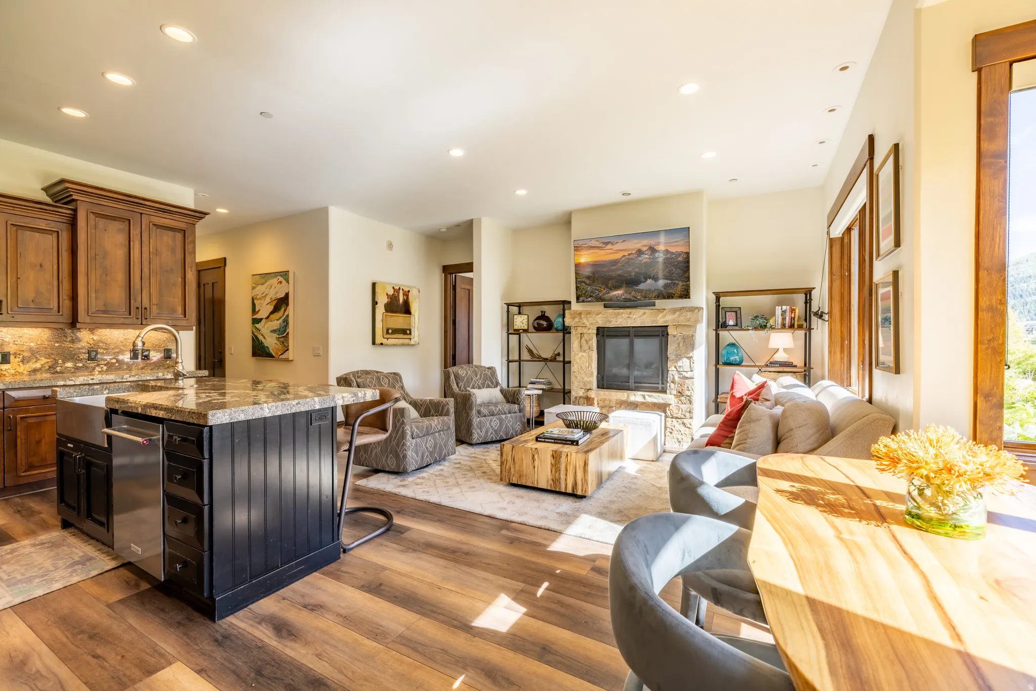 Open-concept living space featuring wood-finish flooring, a stone-surround fireplace, and a kitchen island with built-in appliances and a farmhouse sink