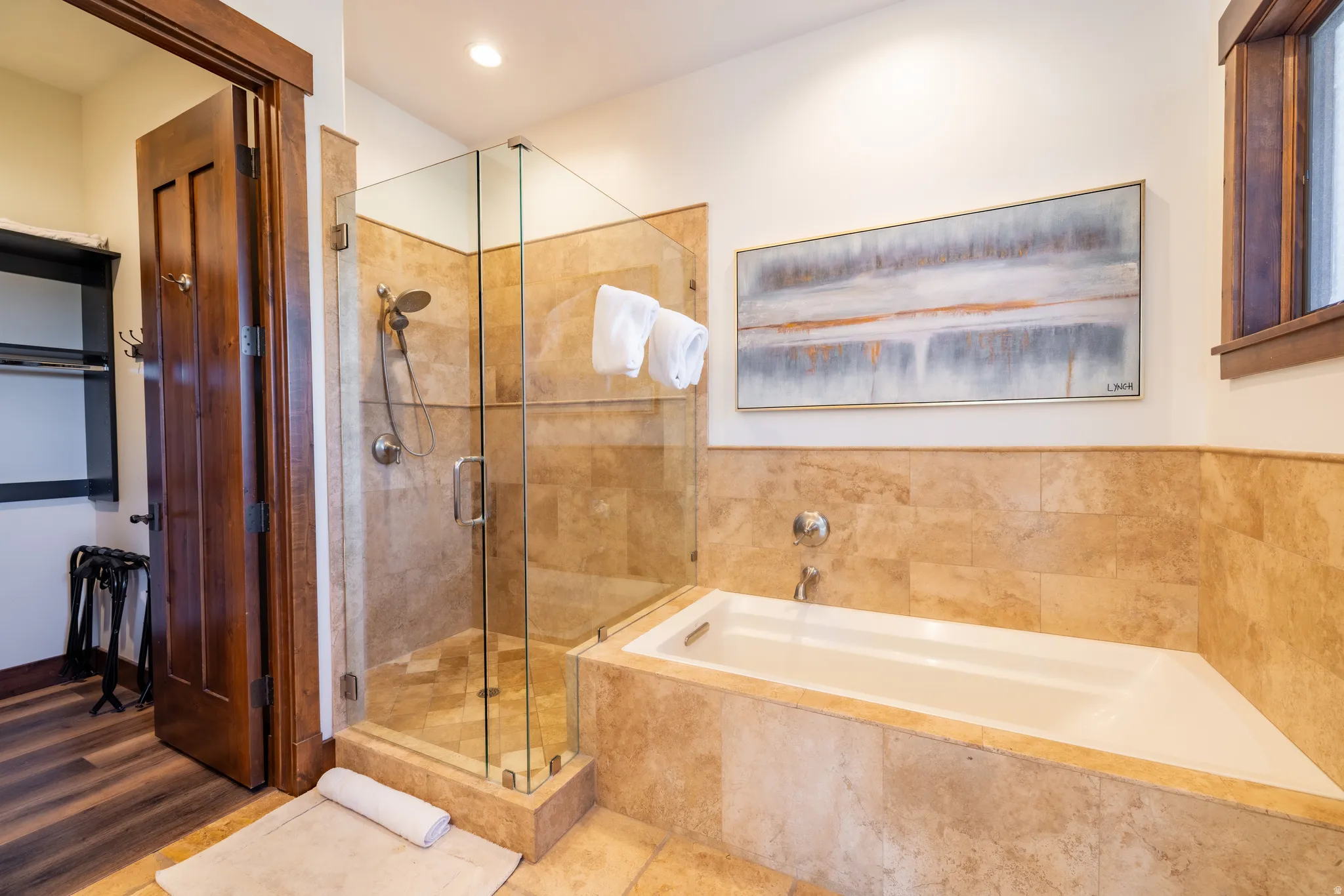 Walk-in shower with clear glass enclosure, a built-in soaking tub, natural stone tile surround, and wood-finish flooring