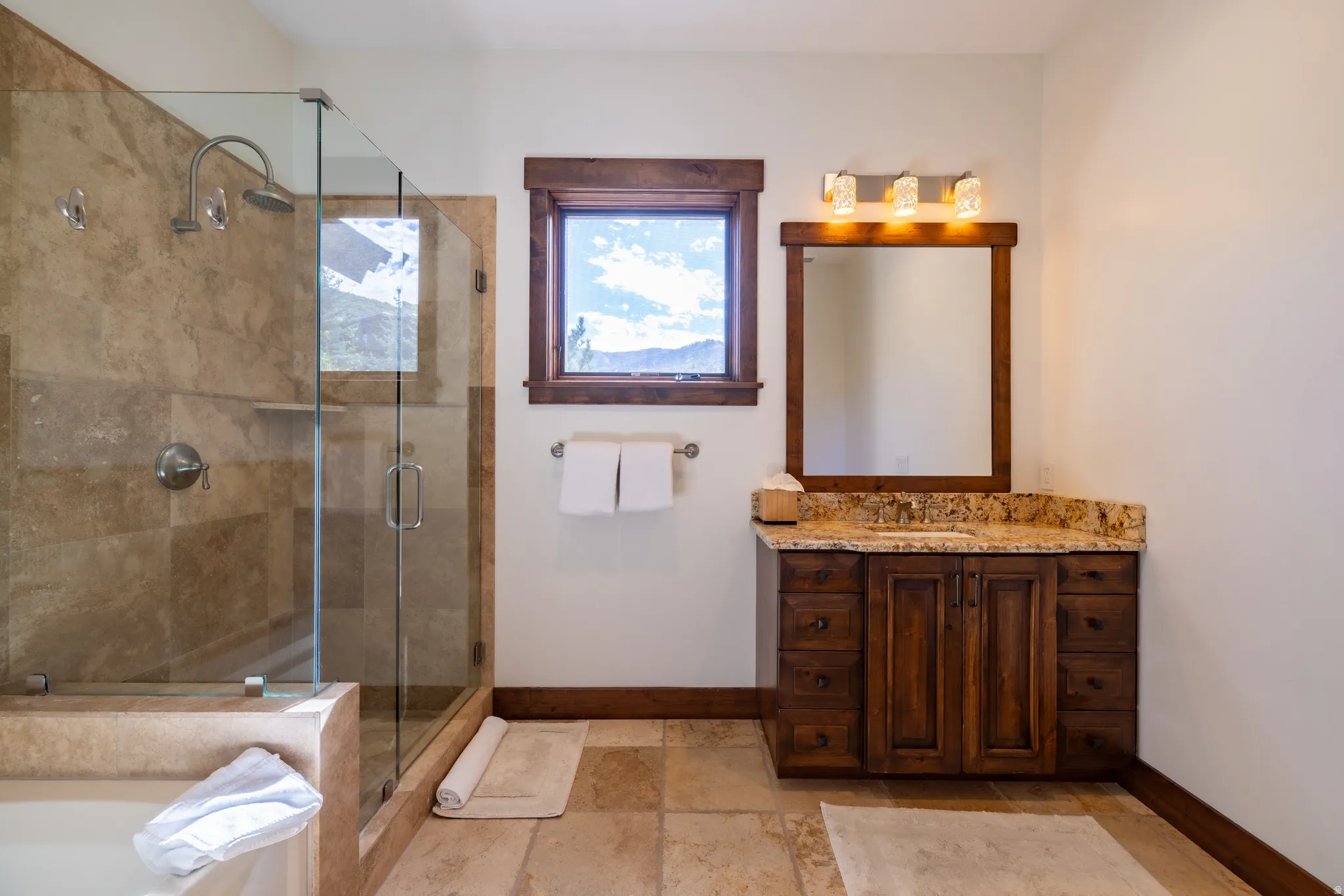 Glass-enclosed shower with stone tile surround, separate soaking tub, wood-finish vanity with stone countertop, and wood-framed mirror
