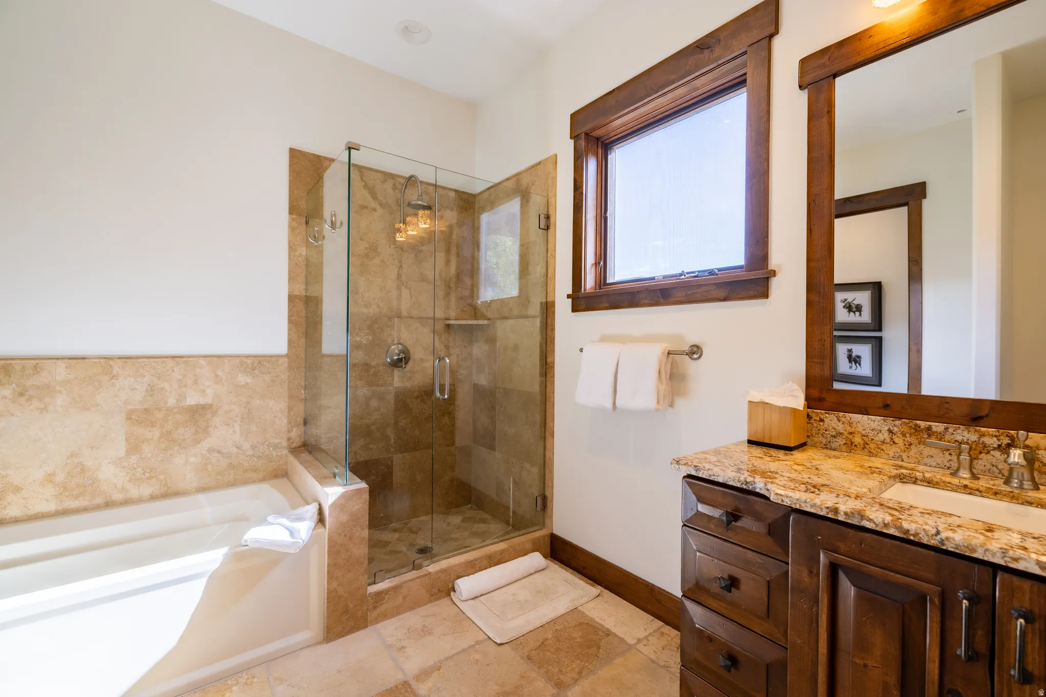 Expansive soaking tub, stone tile shower with glass enclosure, wood-framed window, and a vanity with a stone countertop and integrated sink