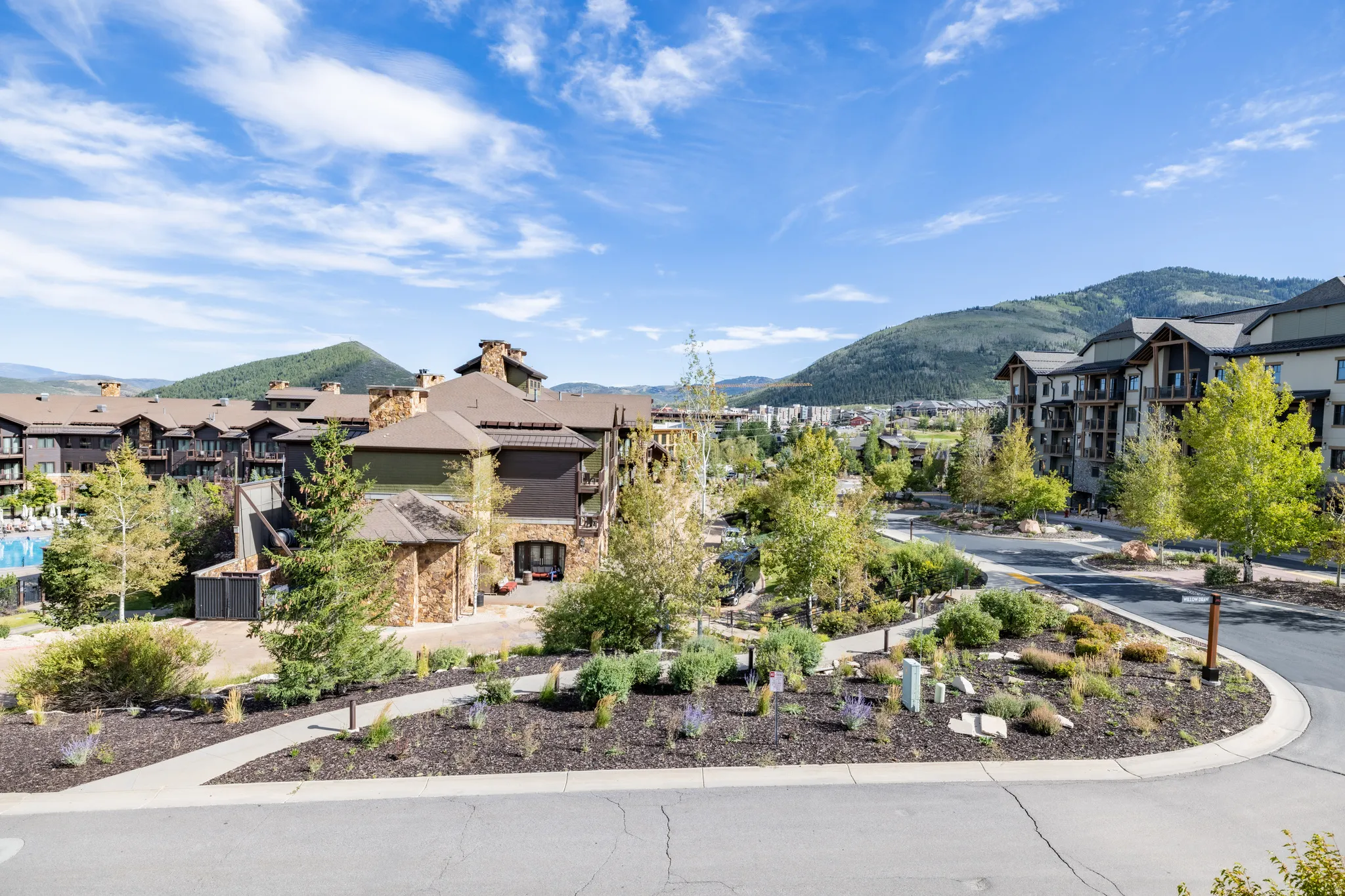 Mountainous terrain backdrop, multiple residential buildings with stone and wood exteriors, manicured landscaping, paved pathways, and a paved road