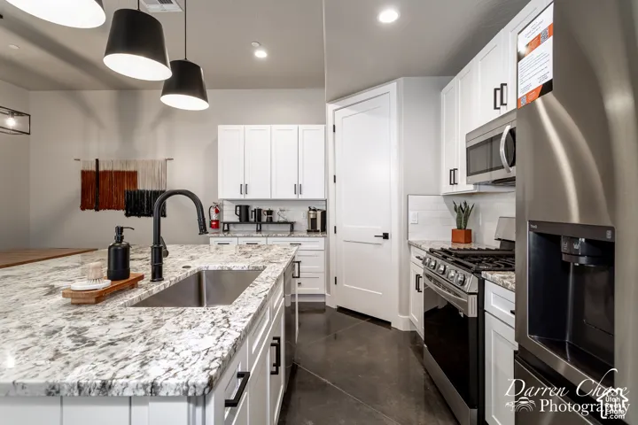 Kitchen featuring white shaker cabinetry, stone countertops, stainless steel appliances, a center island with an undermount sink, and black matte hardware