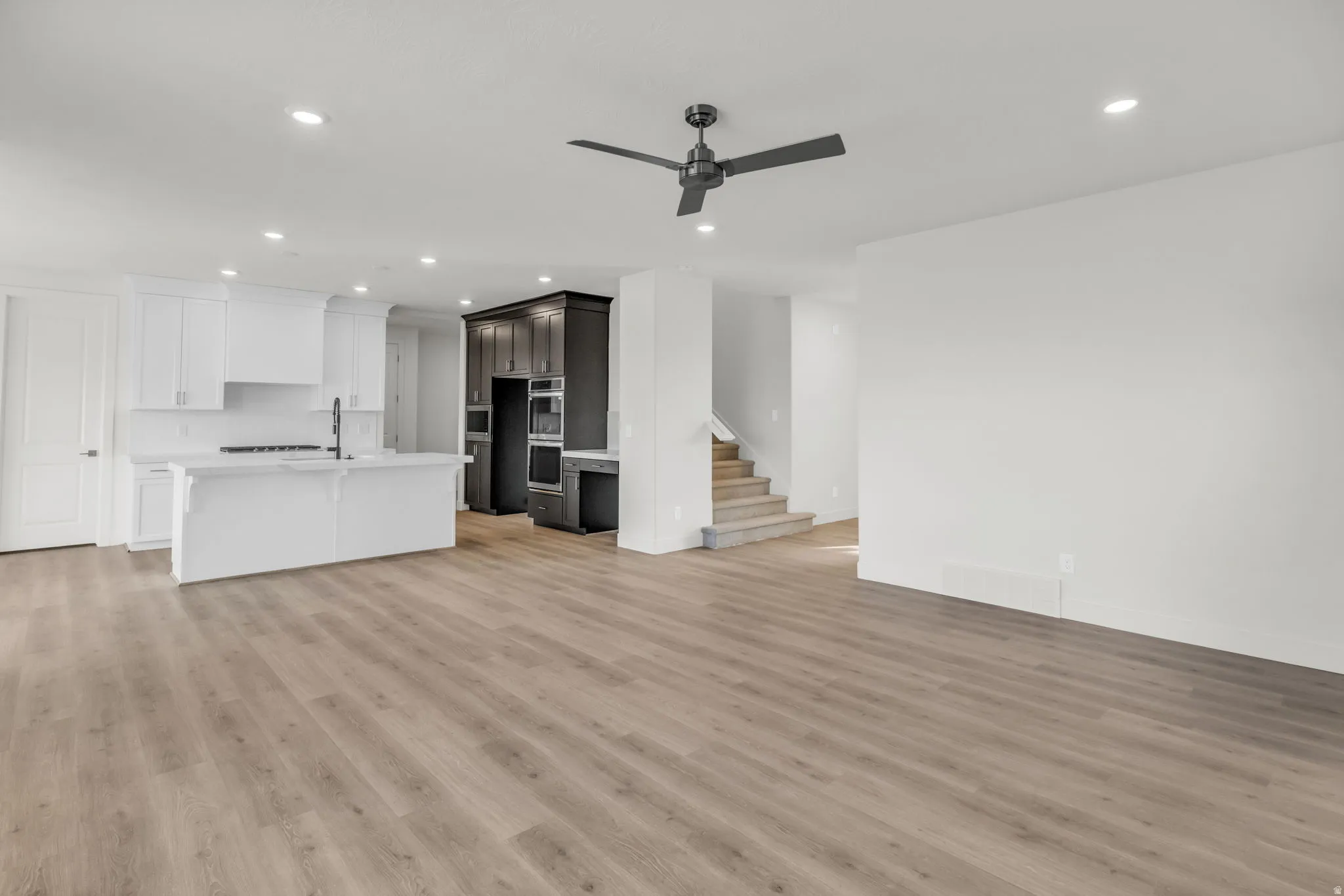 Open-concept living space featuring wood-finish flooring throughout, a modern kitchen with white cabinetry and dark accent shelving, recessed ceiling lighting, a ceiling fan, and a staircase providing access to additional levels