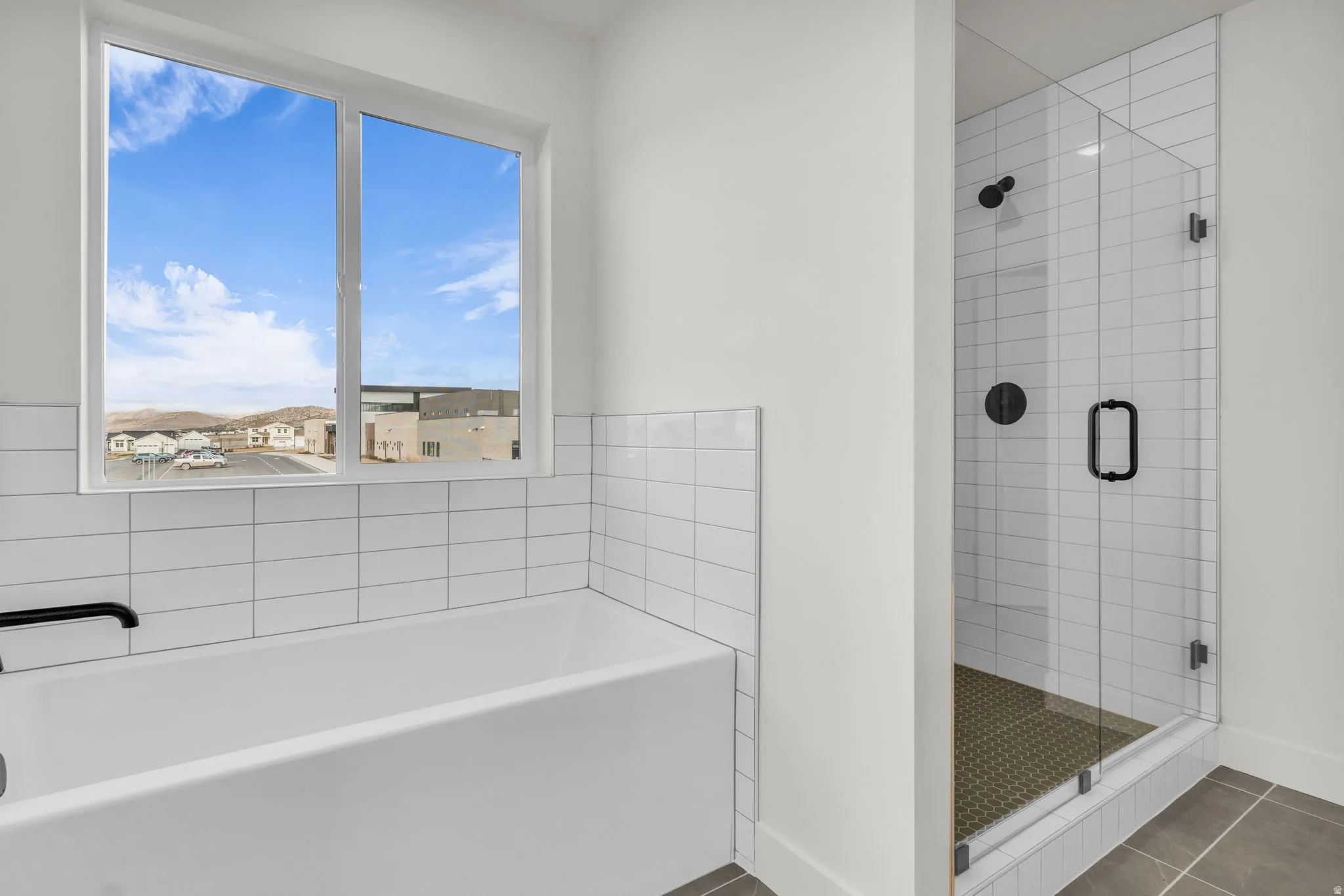 Modern primary bathroom featuring a freestanding soaking tub with subway tile surround, frameless glass walk-in shower enclosure with multiple showerheads and dark tile flooring, white subway tile walls, and a large window with expansive natural light and