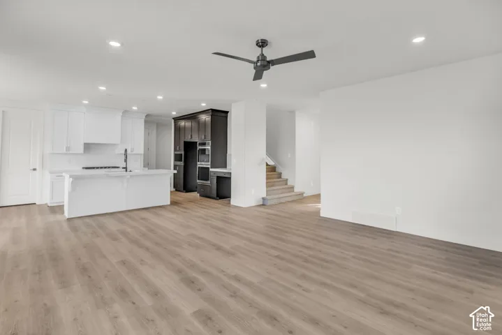 Open-concept living space featuring wood-finish flooring throughout, a modern kitchen with white cabinetry and dark accent shelving, recessed ceiling lighting, a ceiling fan, and a staircase providing access to additional levels
