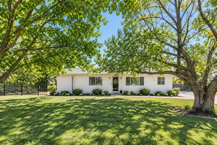 White siding exterior with a dark entry door, multiple windows, established trees, a manicured lawn, and dark metal fencing