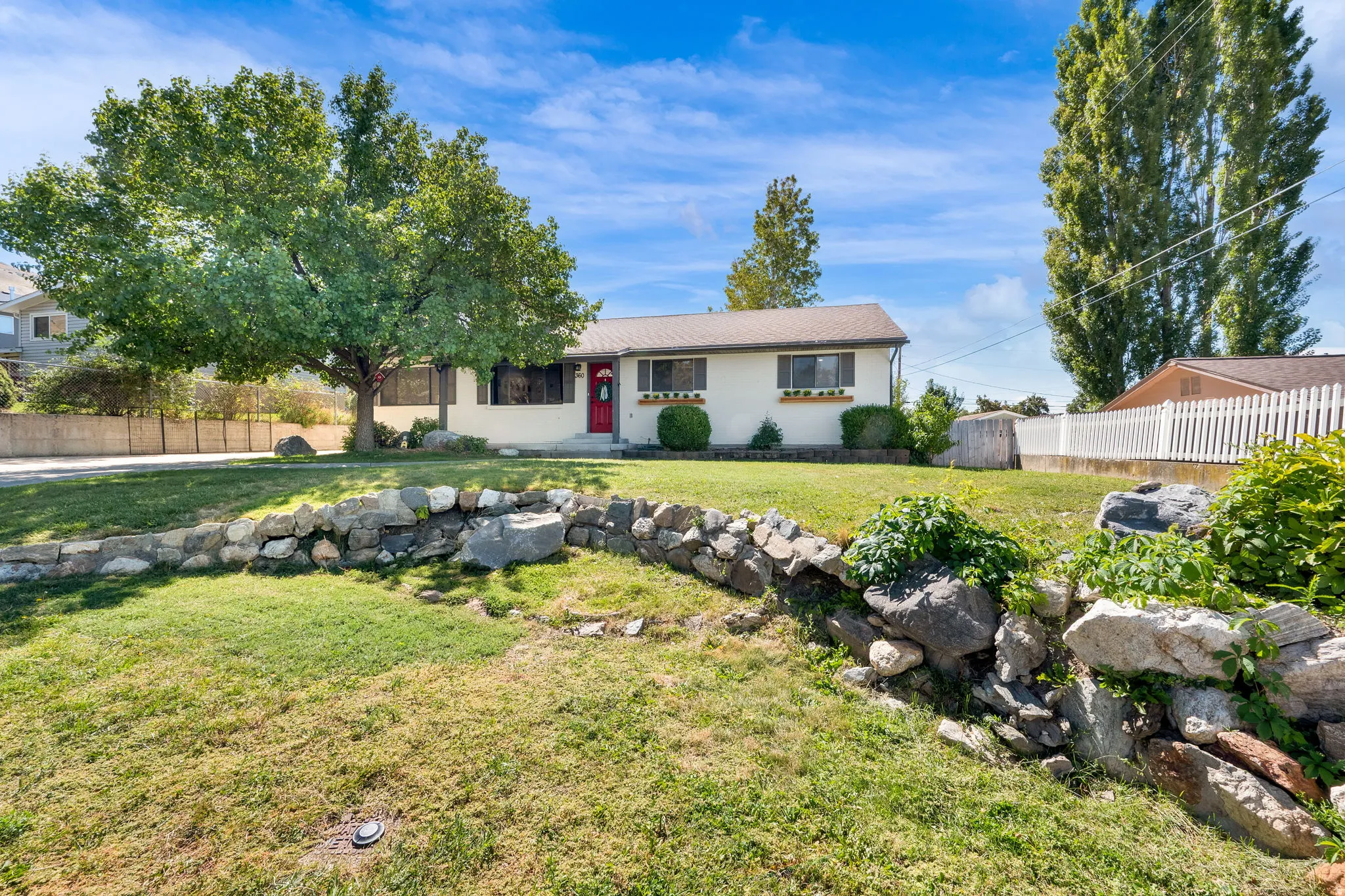 Single-story residence featuring a red entry door, front lawn with mature trees, and a stone retaining wall