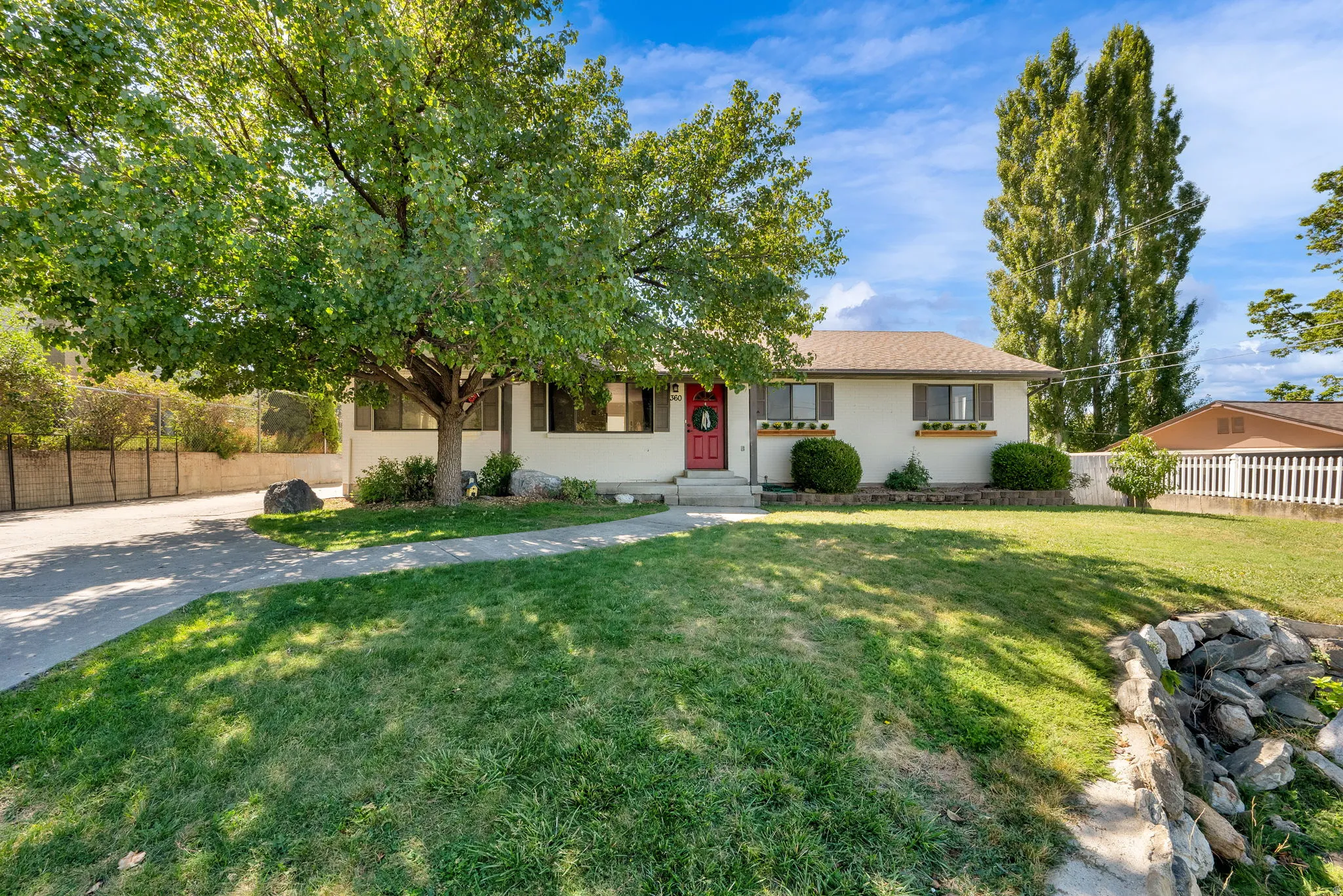 Front exterior featuring a red entry door, decorative window boxes, mature front yard tree, established shrubbery, and a paved driveway