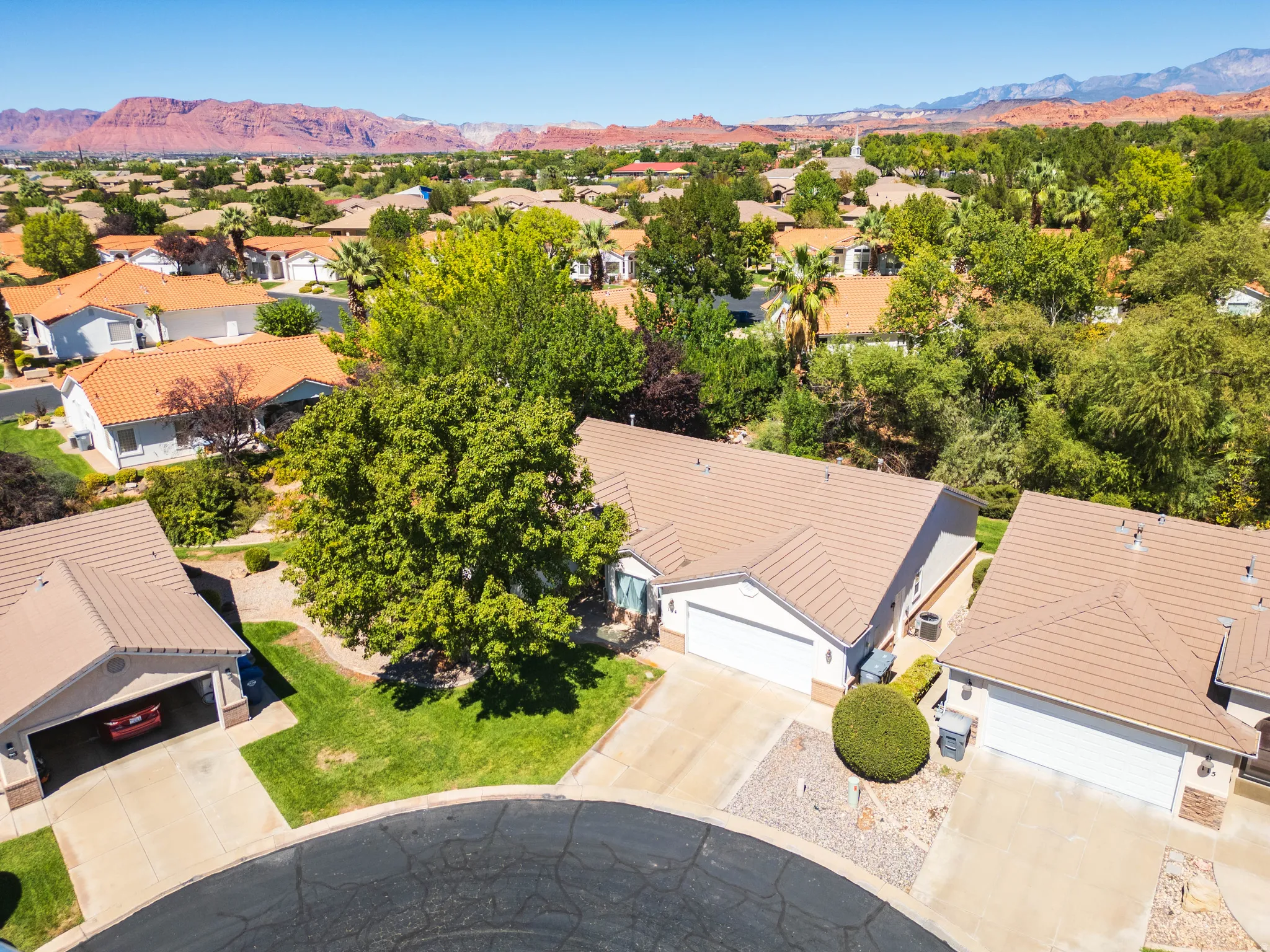 Aerial perspective of a neighborhood with red rock mountain backdrop, featuring homes with tile roofs, mature trees, and landscaped yards