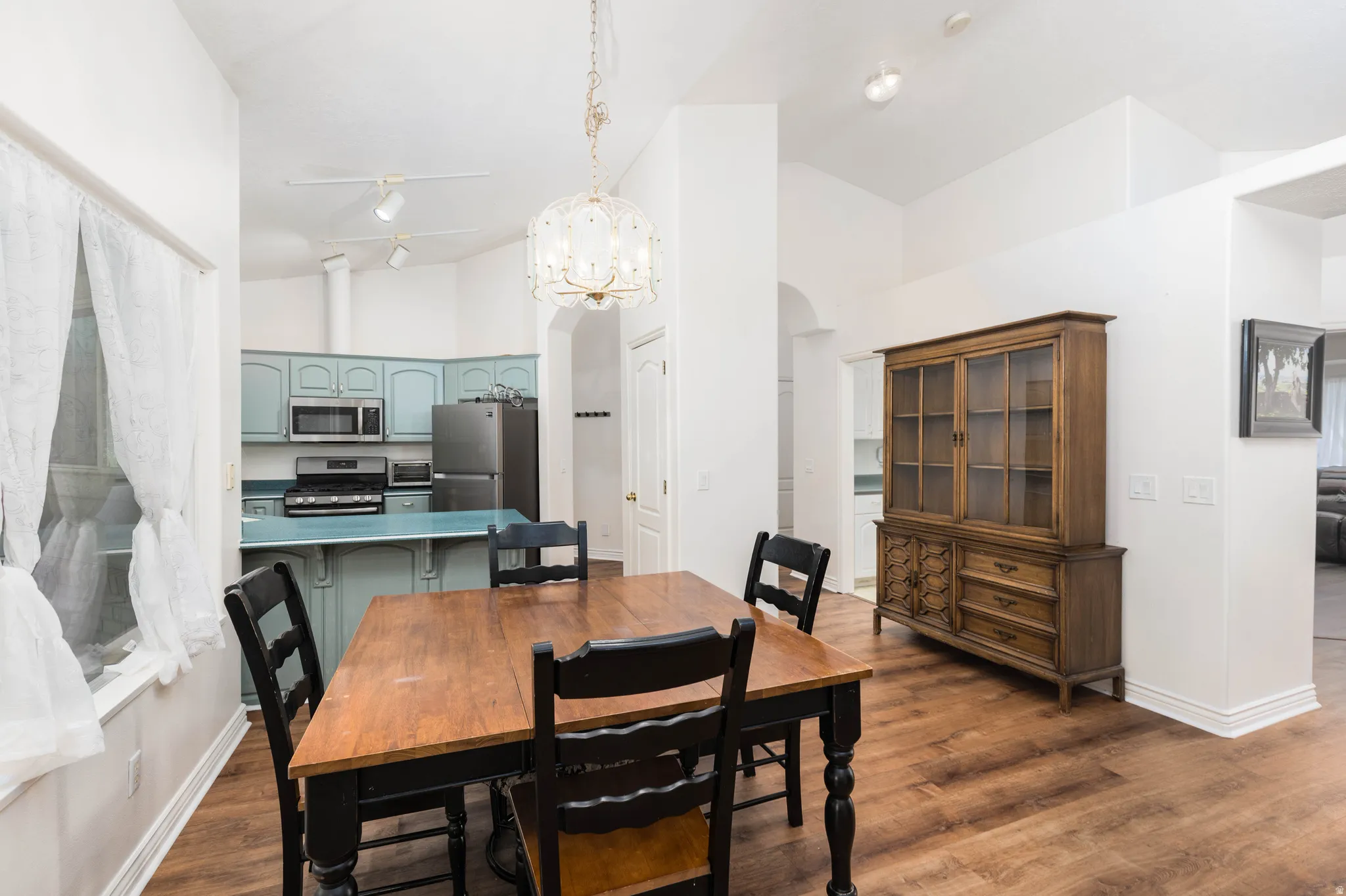 Vaulted ceilings with track lighting, wood-finish flooring, kitchen with light blue cabinetry and stainless steel appliances, and a built-in pantry door