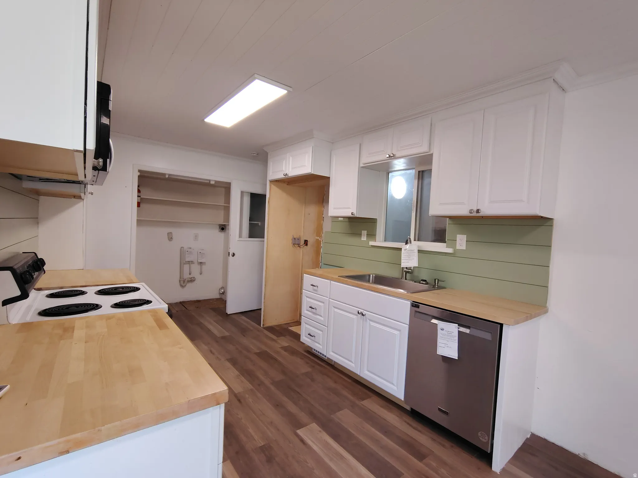 Kitchen with wood-finish flooring, white cabinetry, light wood countertops, stainless steel dishwasher, and a green shiplap backsplash