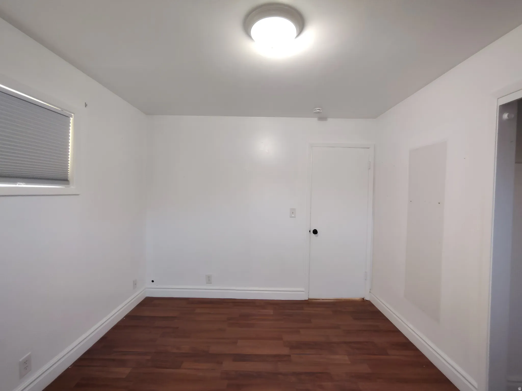 Wood-finish flooring with white baseboards, a single window with blinds, and a flush mount ceiling light