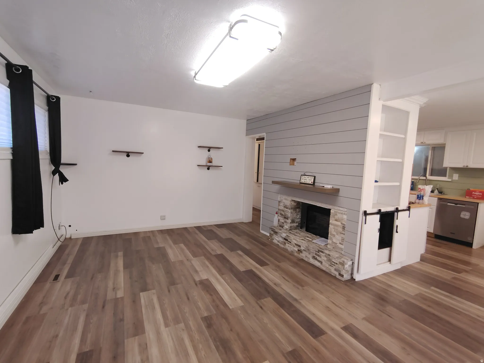 Wood-finish flooring, stone-clad fireplace with wood mantle, shiplap accent wall, built-in shelving, and stainless steel dishwasher