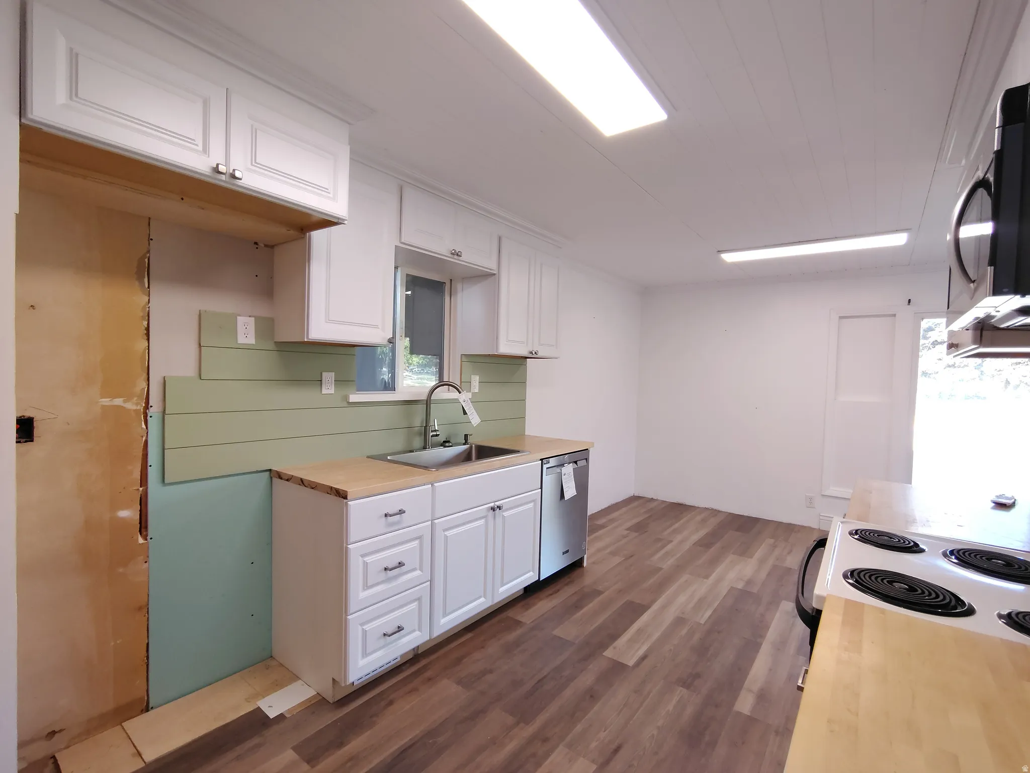 Galley kitchen featuring white cabinetry with multiple drawers and storage, stainless steel sink and dishwasher, sage green subway tile backsplash, wood-finish countertops, and wood-look plank flooring throughout