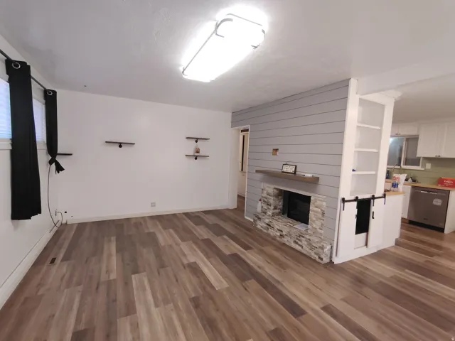 Wood-finish flooring, stone-clad fireplace with wood mantle, shiplap accent wall, built-in shelving, and stainless steel dishwasher