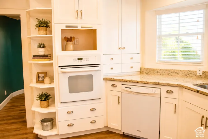 White cabinetry with brass hardware, light-toned stone-finish countertops, built-in wall oven and microwave, corner open shelving, and wood-finish flooring