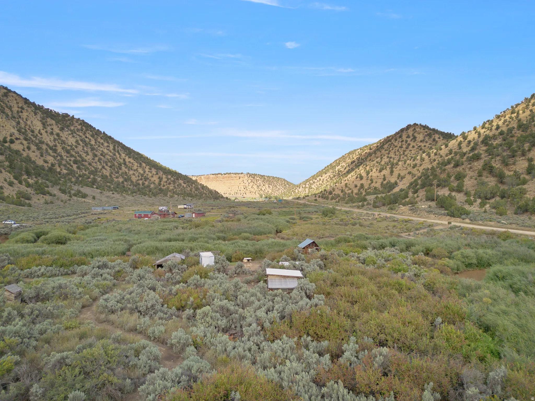 Expansive property with natural vegetation, framed by mountainous terrain, featuring distant structures, and a visible unpaved road