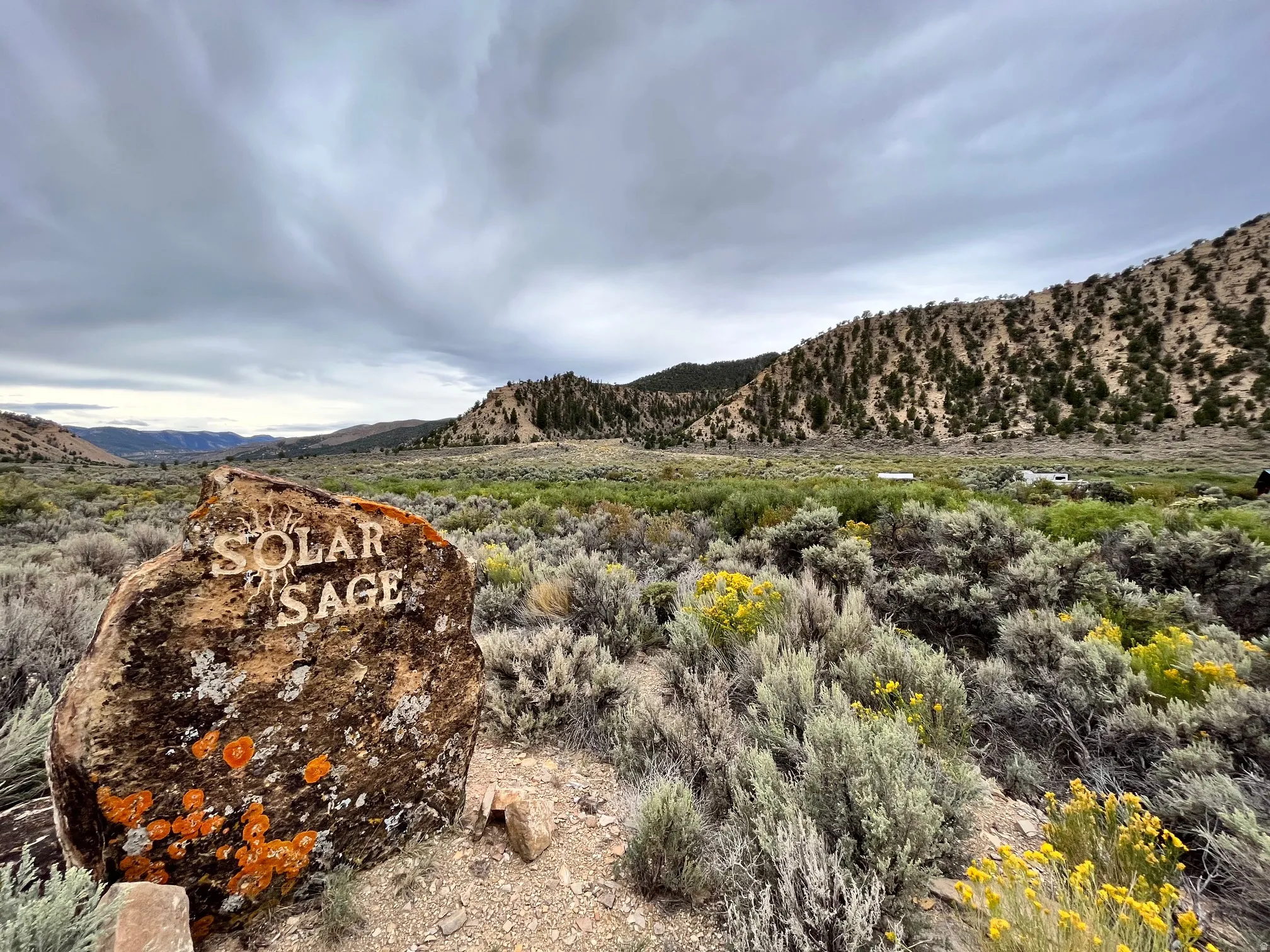 Expansive natural landscape featuring a prominent rock with "SOLAR SAGE" inscription, diverse shrubbery, and distant tree-covered hills
