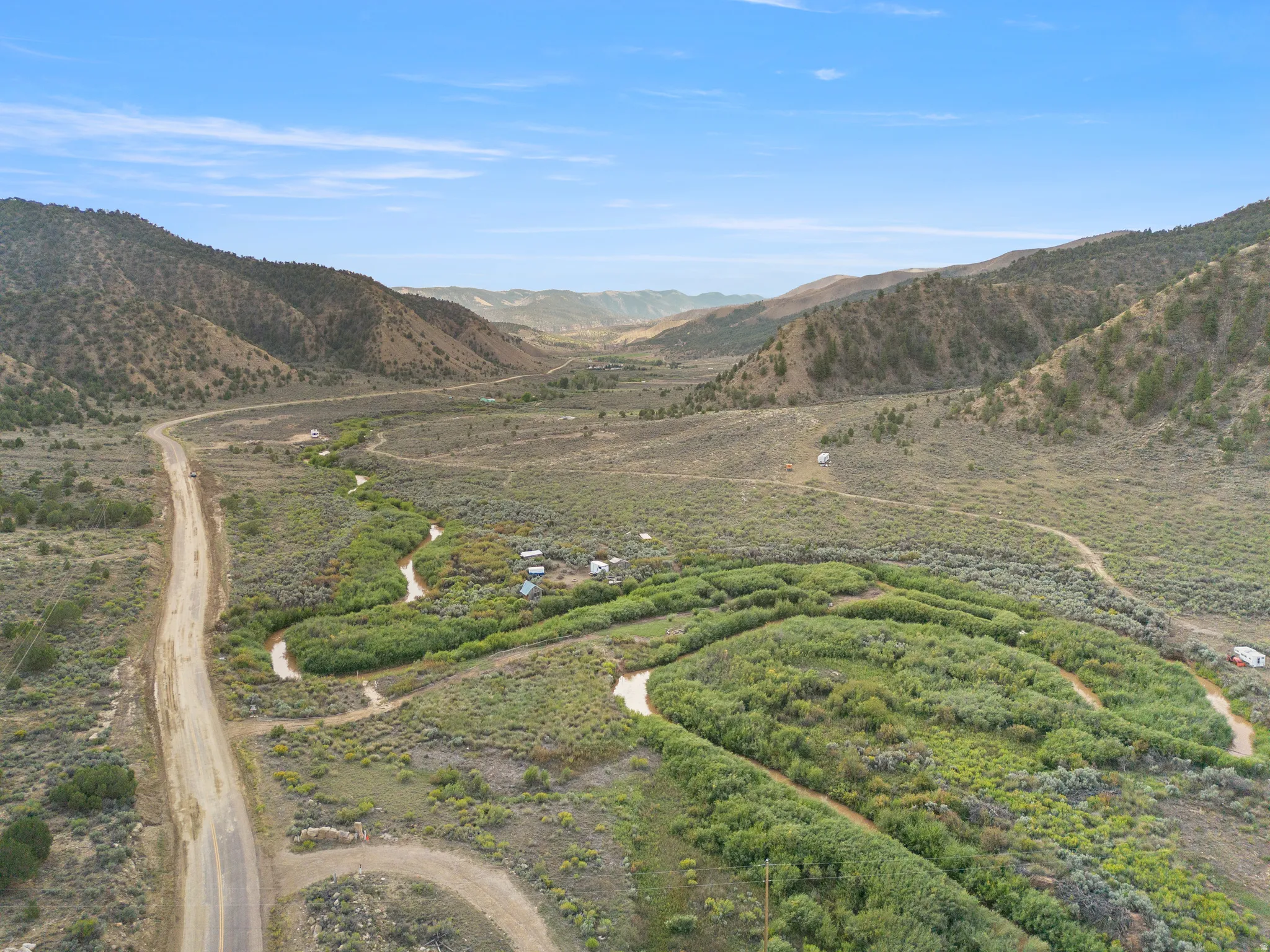 Expansive rural landscape featuring a winding dirt road, natural watercourse with riparian vegetation, and mountainous terrain