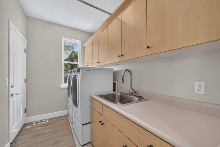 Laundry area featuring wood-finish flooring, ample cabinetry, a utility sink with a gooseneck faucet, and a large window