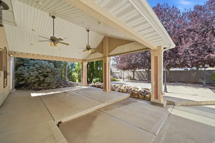 Expansive covered patio featuring a vaulted ceiling with two ceiling fans, substantial support columns, concrete flooring, and surrounding mature trees