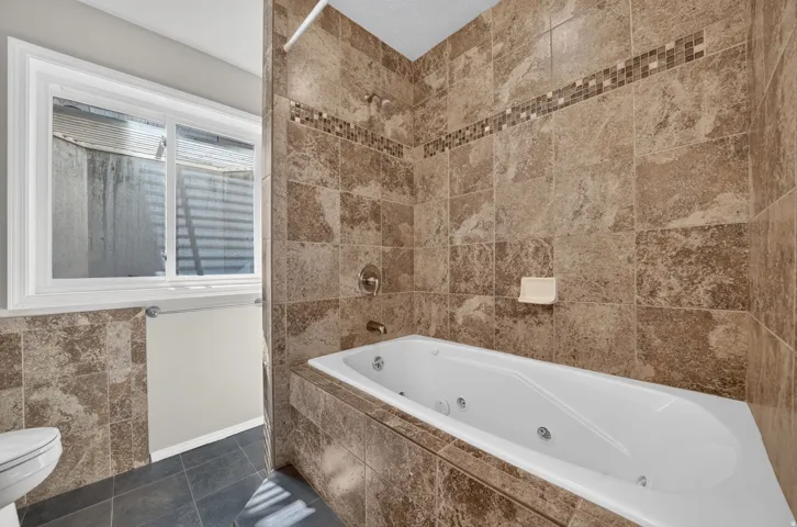 Jetted bathtub with full-height tile surround, mosaic tile accent band, and a window for natural light