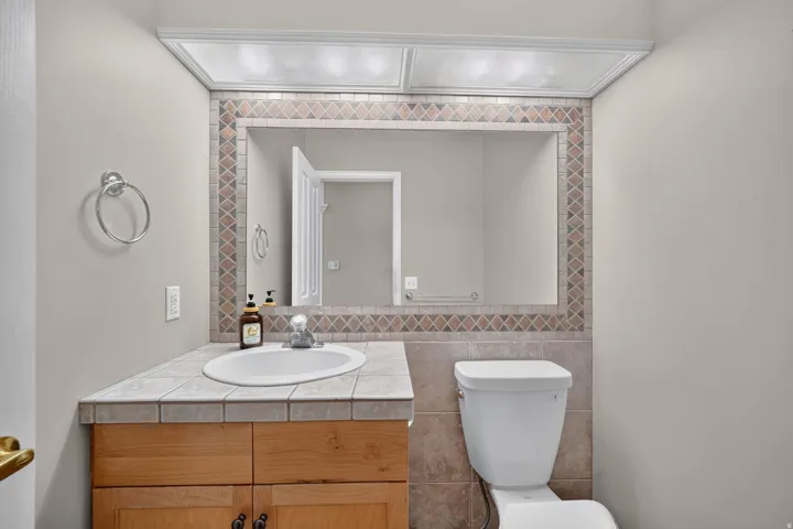 Bathroom featuring a wood-finish vanity with tile countertop, an undermount sink, a tiled backsplash, and a mirrored wall